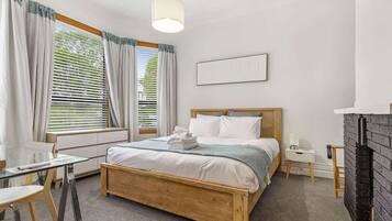 A bedroom with a wooden bed, a glass table, a white bedside table, a fireplace, and a window with curtains.