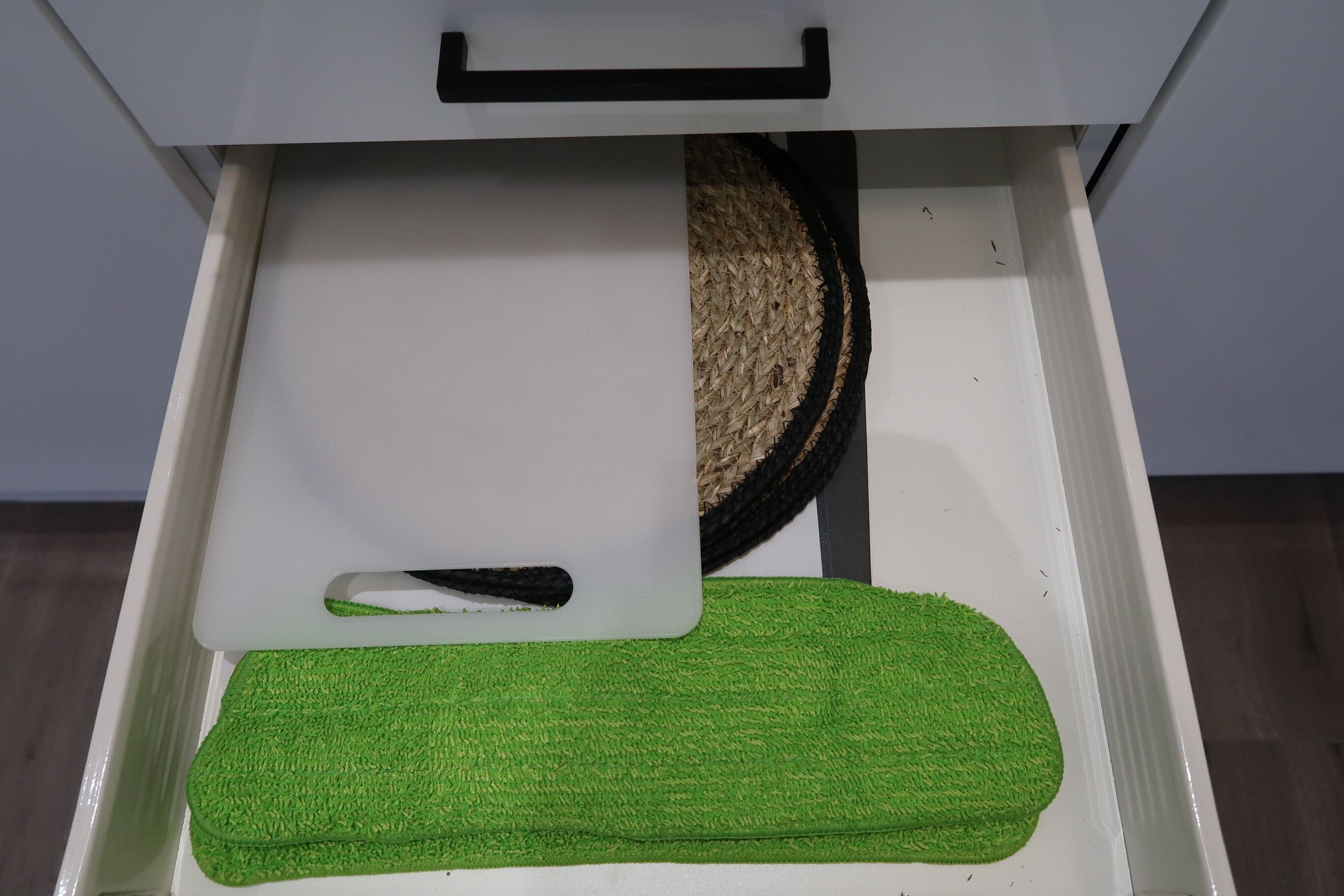 Een witte lade met een zwarte handgreep, waarin een wit voorwerp en een grof bewerkte, groene mat liggen.