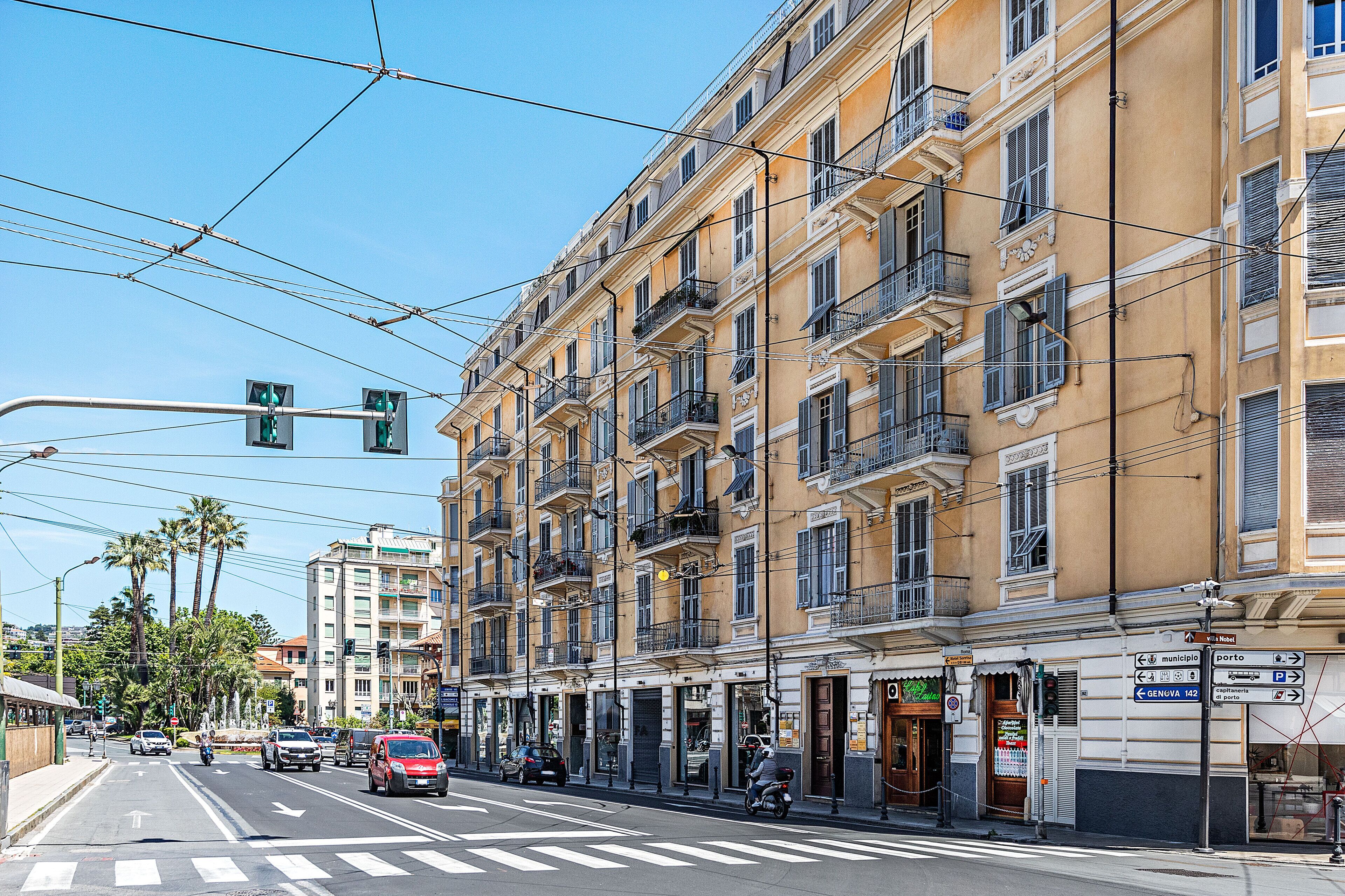 Un edificio a più piani con balconi, una strada con auto e un attraversamento pedonale, e linee elettriche aeree.