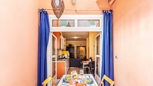 A dining area with a table set for two, a kitchen in the background, and blue curtains on the windows. - Gaudio Charming Apartment by Wonderful Italy (Sanremo)