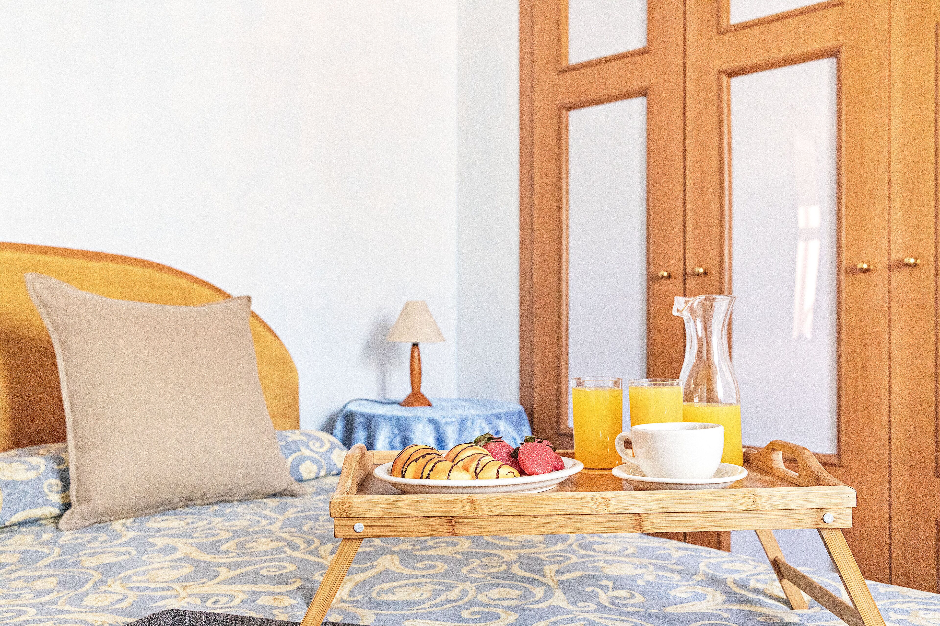 Una cama con cabecera, una mesita de noche con lámpara, una bandeja con pasteles y un vaso de jugo de naranja, y una puerta de madera al fondo.