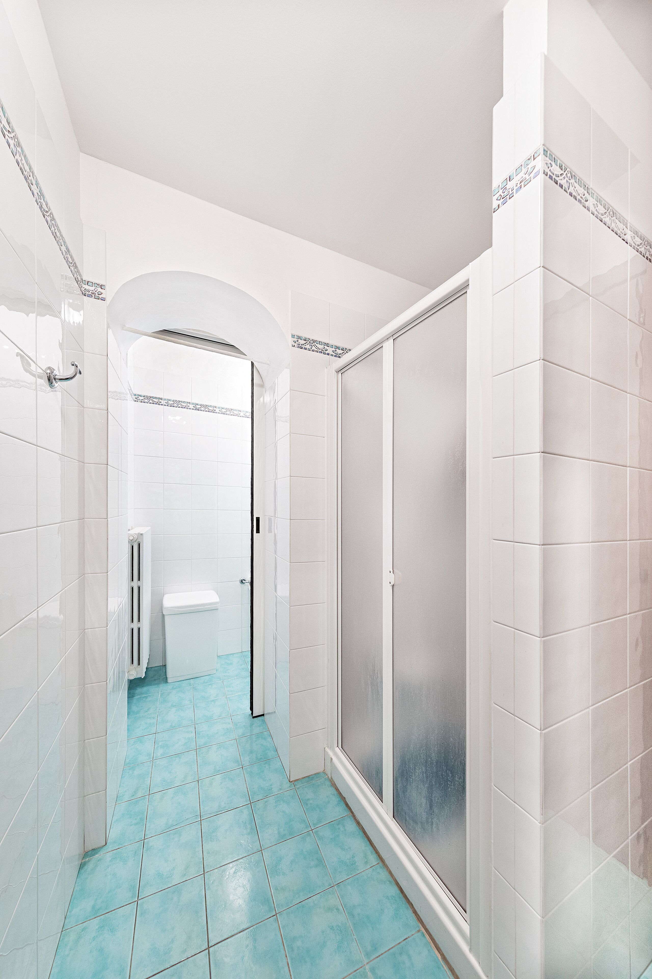 Une salle de bain avec des murs recouverts de carreaux blancs, une douche dotée d’une porte en verre et un sol carrelé de bleu.