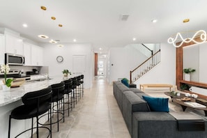 A modern kitchen with a marble countertop, black bar stools, and a dining area with a grey sofa and a wooden coffee table.