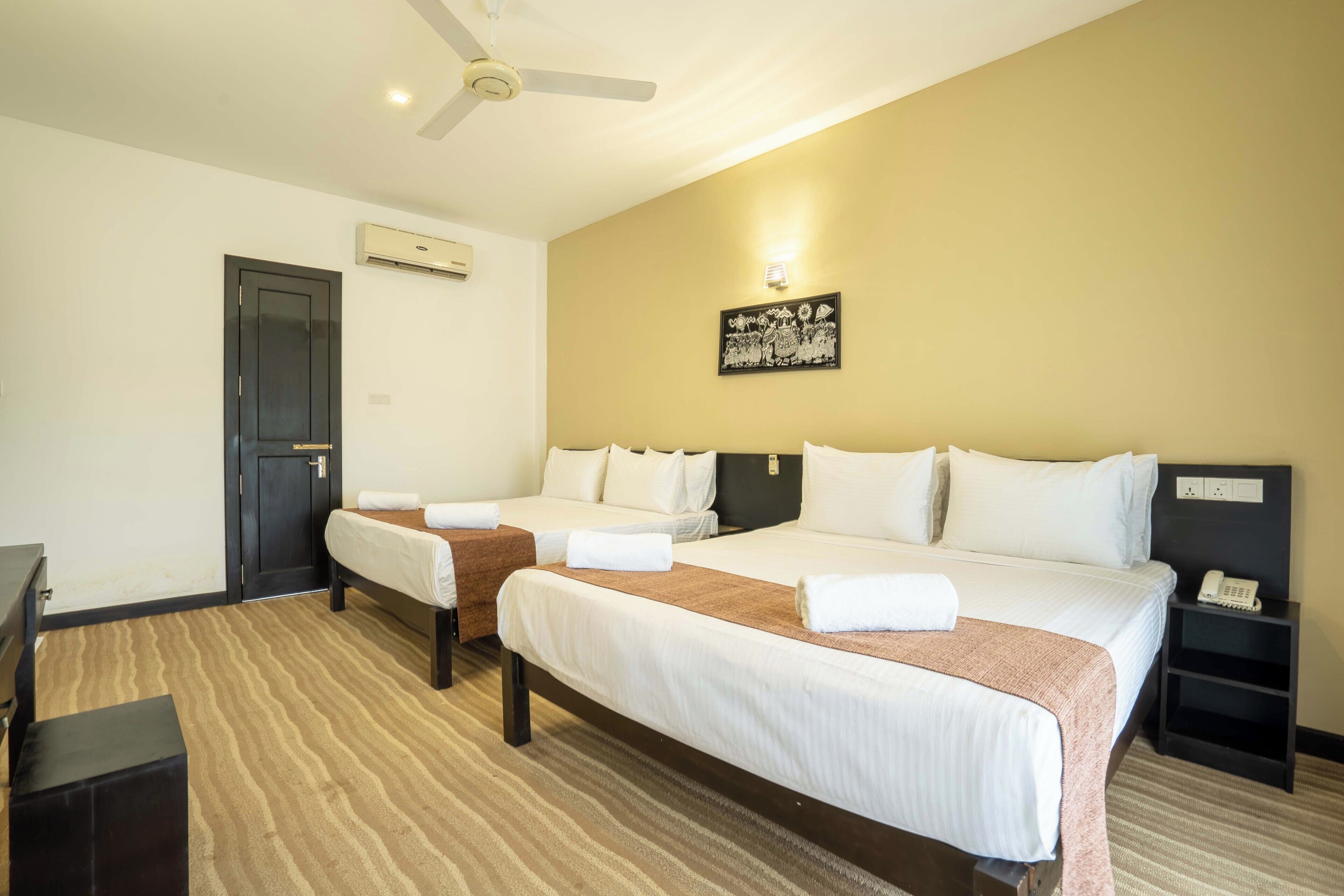 A hotel room with two beds, a ceiling fan, an air conditioning unit, a wall-mounted artwork, and a bedside telephone.