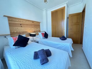 Habitació d'hotel amb dos llits, un panell de fusta al capçal, una tauleta de nit amb una làmpada i una porta.