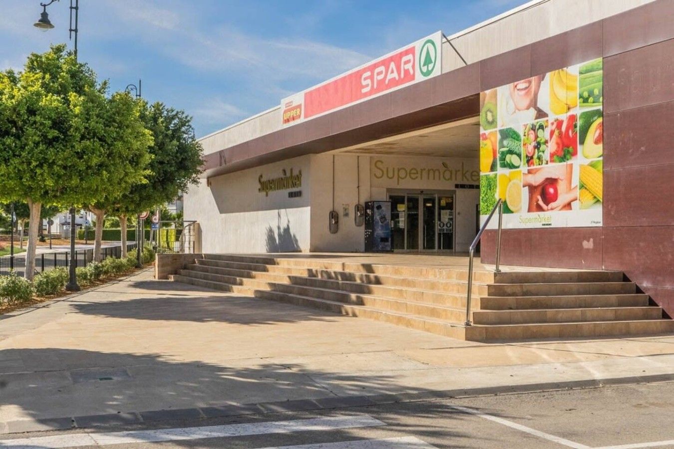 L'ingresso di un supermercato con una rampa che conduce alla porta, un cartello sopra l'ingresso e una grande pubblicità sul lato destro.