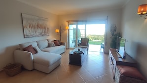 A living room with a sofa, coffee table, and a television. There is a sliding glass door leading to an outdoor area with a chair and a table. - Sand, Sea and Salt - Alvor (Alvor)