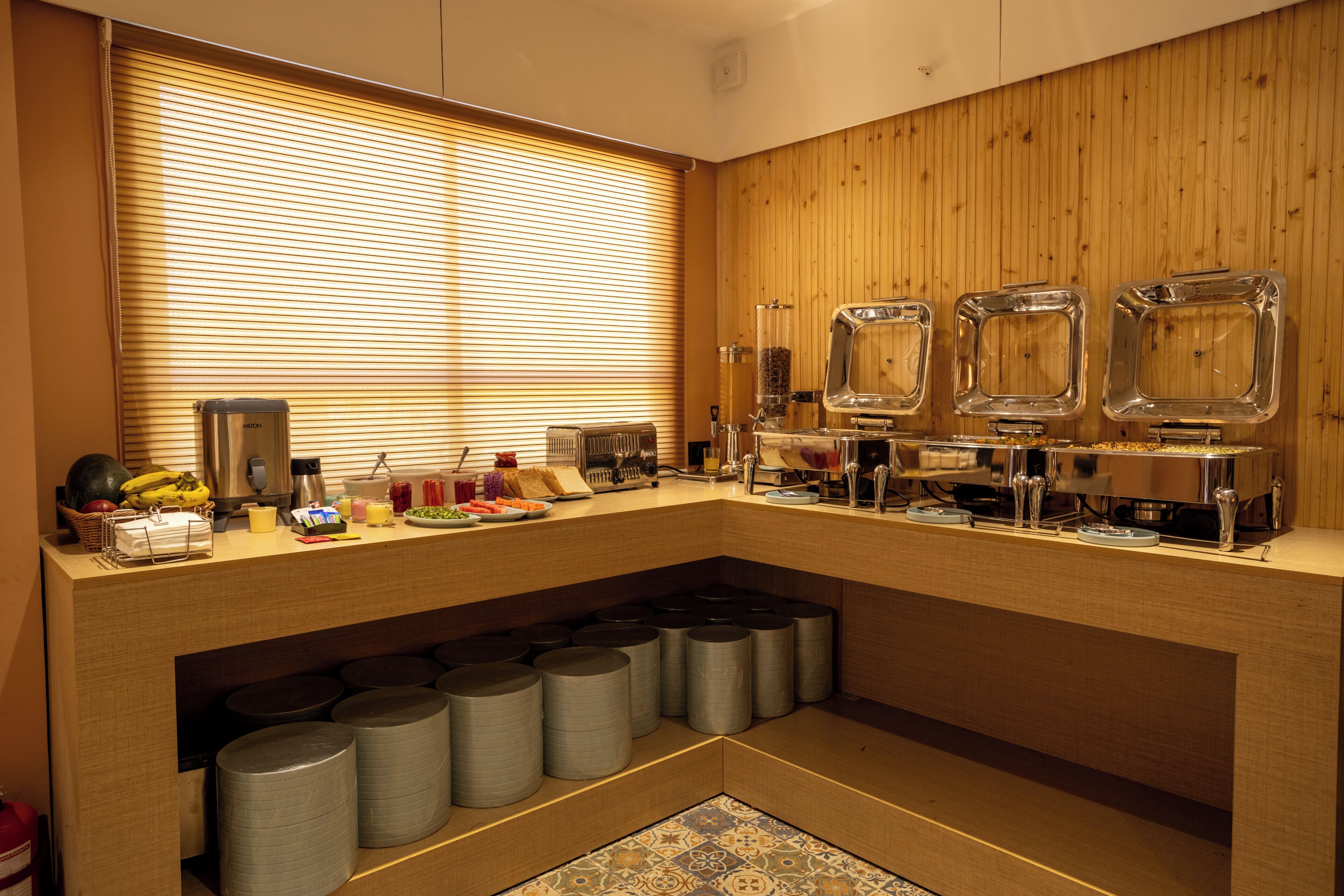 A breakfast bar with a wooden backdrop, a window with blinds, and various breakfast items.