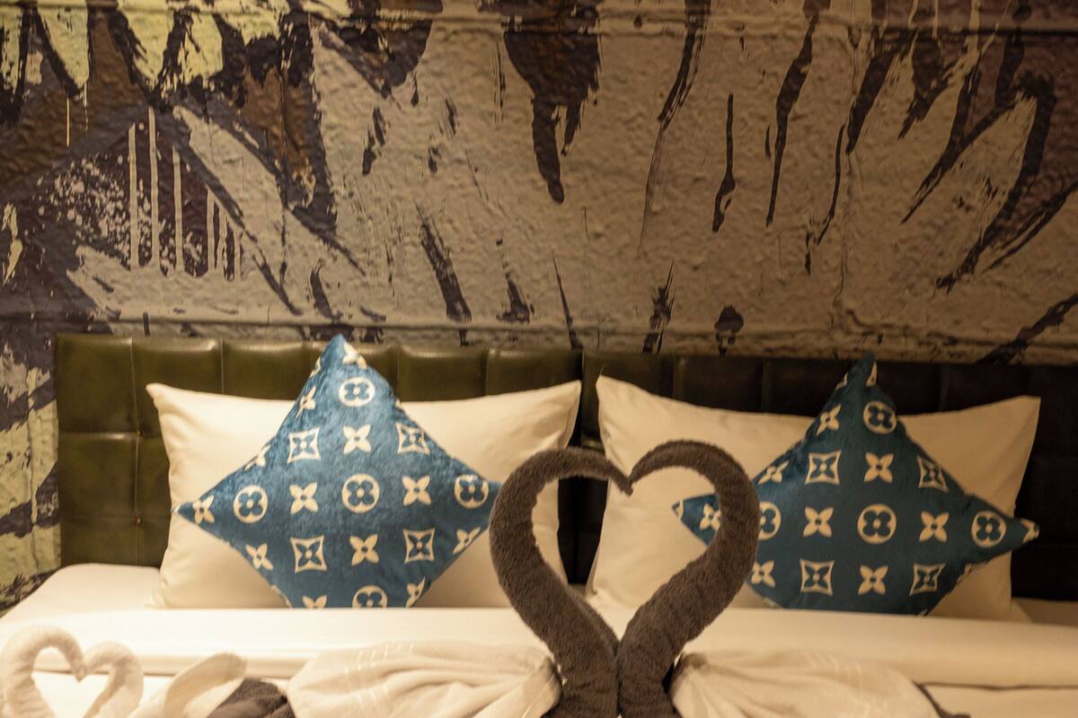 A bed with a blue patterned bedspread, two pillows with a monogram design, and a heart-shaped towel arrangement.