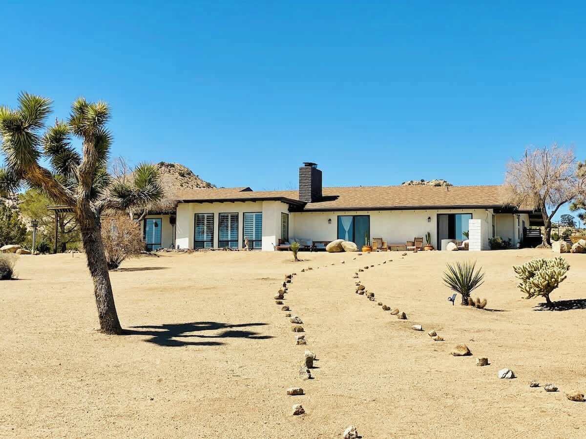 Une maison de plain-pied, entourée d’un paysage désertique, avec un ciel dégagé et un chemin menant à l’entrée.