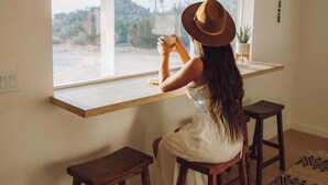 A person sitting at a wooden table by a large window, with a view of a landscape outside. - Villa Paloma • 4-Acre Stylish Retreat with Hot Tub (Yucca Valley)