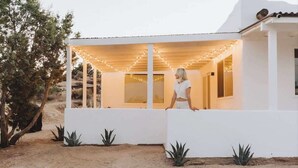 A white building with a covered patio area, surrounded by desert vegetation. - Villa Paloma • 4-Acre Stylish Retreat with Hot Tub (Yucca Valley)