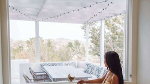 A person sitting on a white sofa with patterned cushions, near a large window with a view of outdoor furniture and trees. - Villa Paloma • 4-Acre Stylish Retreat with Hot Tub (Yucca Valley)