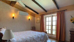 A bedroom with a bed, a nightstand, a lamp, an air conditioning unit, a window with curtains, and a wooden ceiling. - Chambre D'hôtes et Petit Déjeuner D'exceptionnel (Tourrettes)