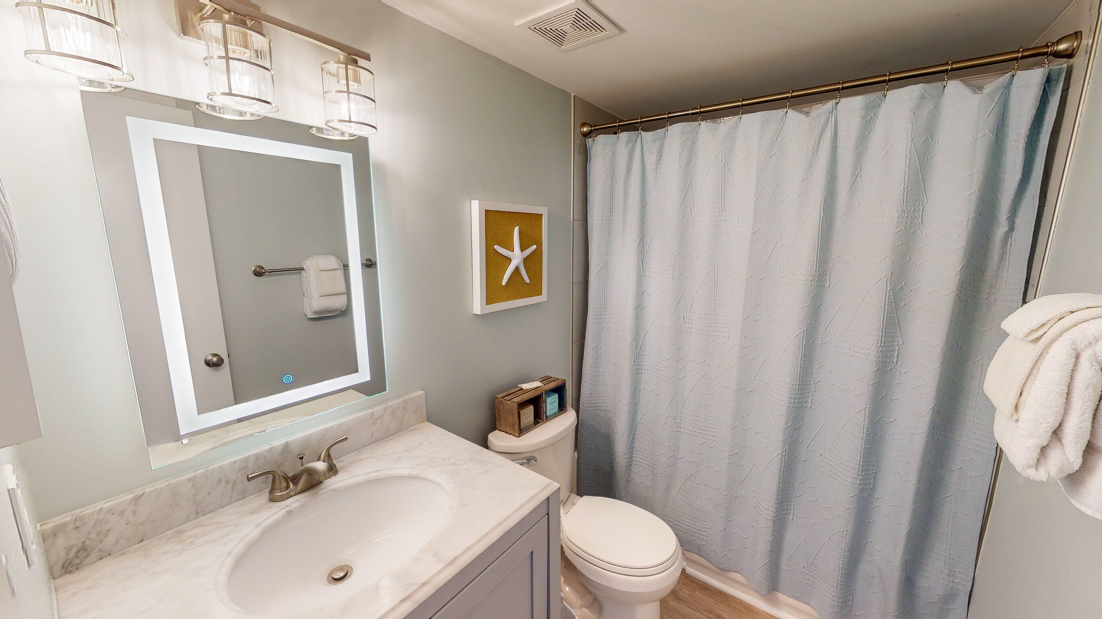 A bathroom with a marble sink, a toilet, a shower curtain, and a mirror with lights.