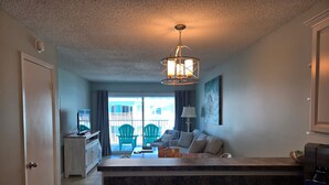 A modern kitchen with a central island, a dining area with chairs, a TV, and a view of the outside. - Top Floor Beachside Condo in Gulf Shores Sleeps 6 (Gulf Shores)