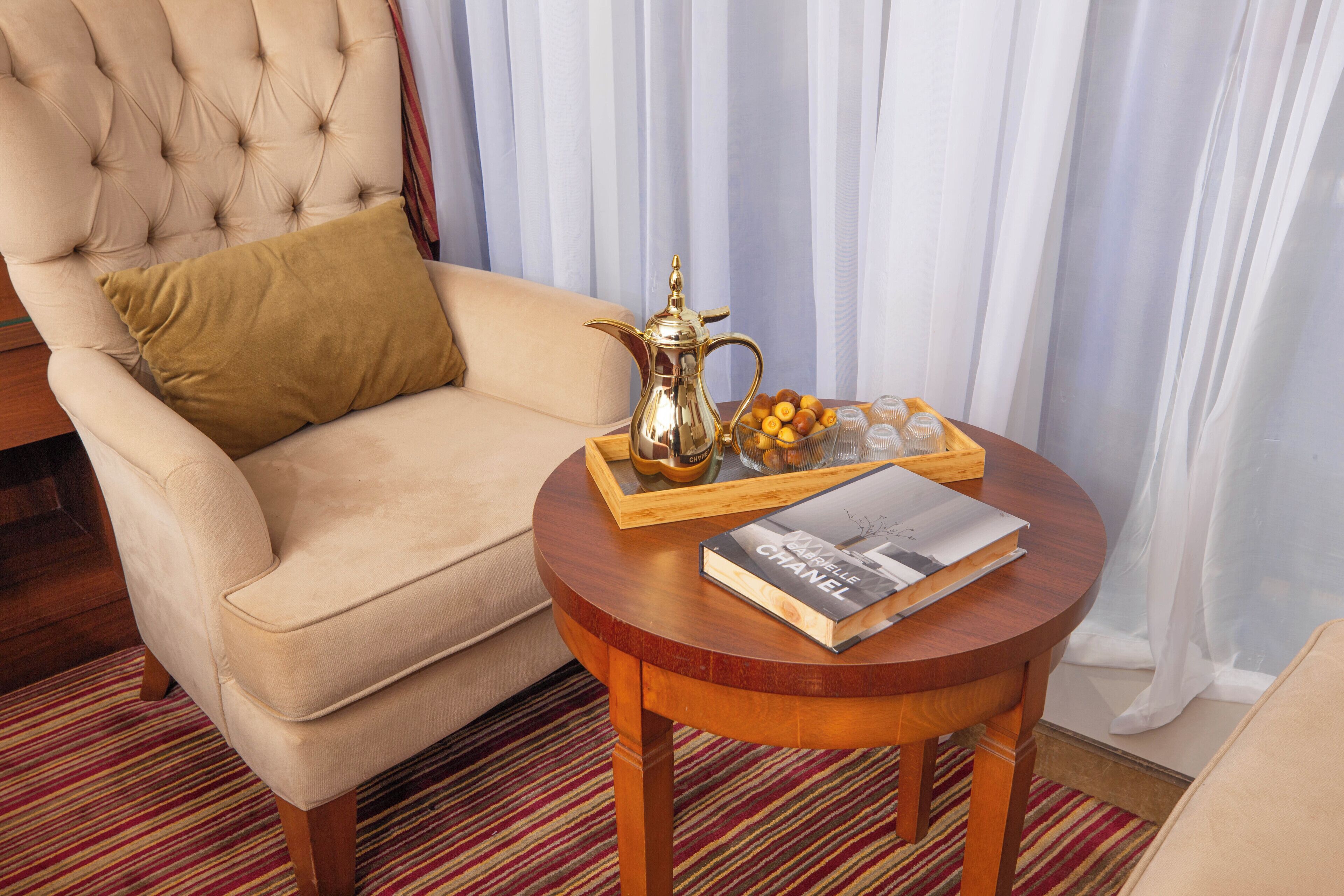Una habitación con un sillón capitoné, una mesa redonda de madera con una bandeja de frutas, un libro y una jarrón decorativo.