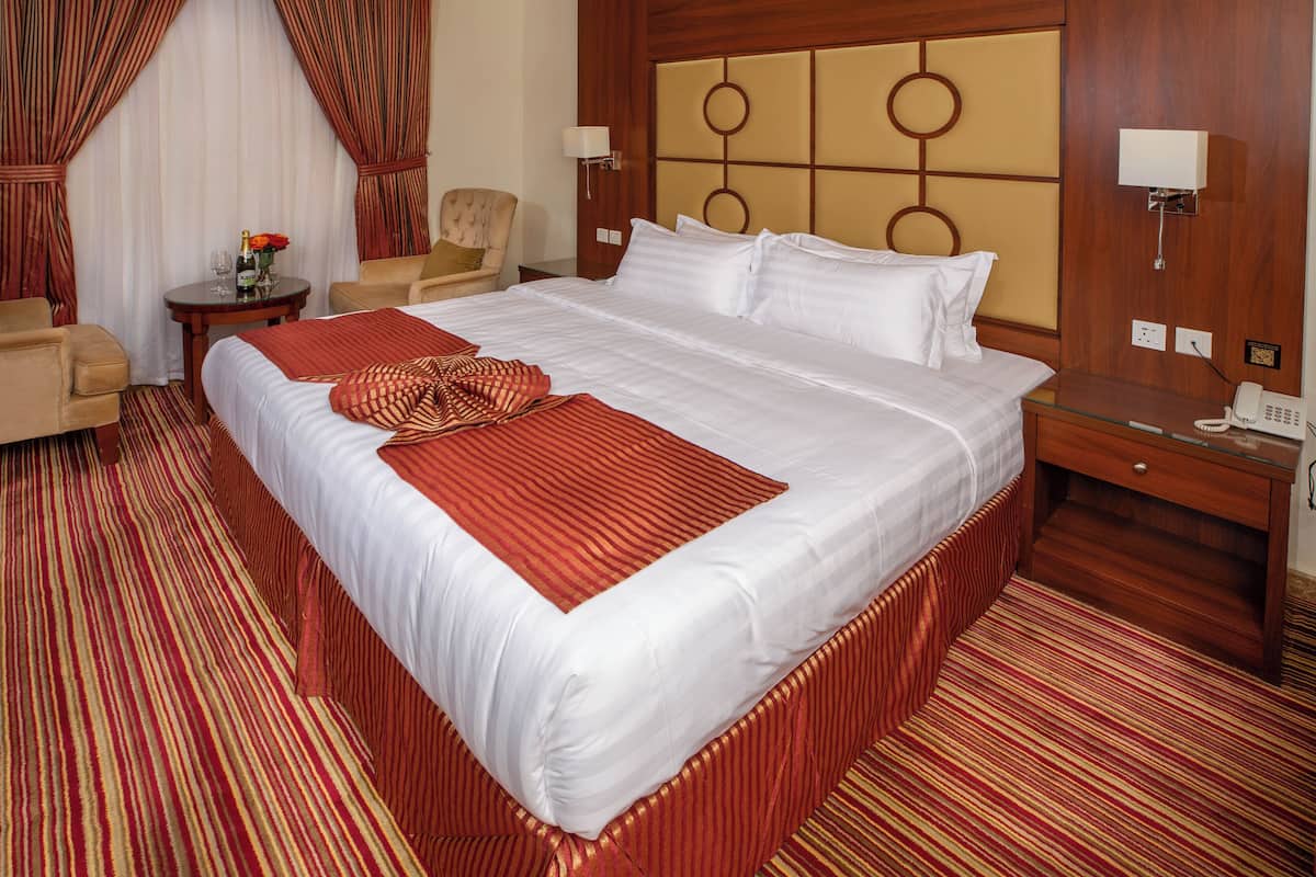 Una habitación de hotel con una cama grande, una silla, una mesita con una botella y vasos, y una mesita de noche con un teléfono.