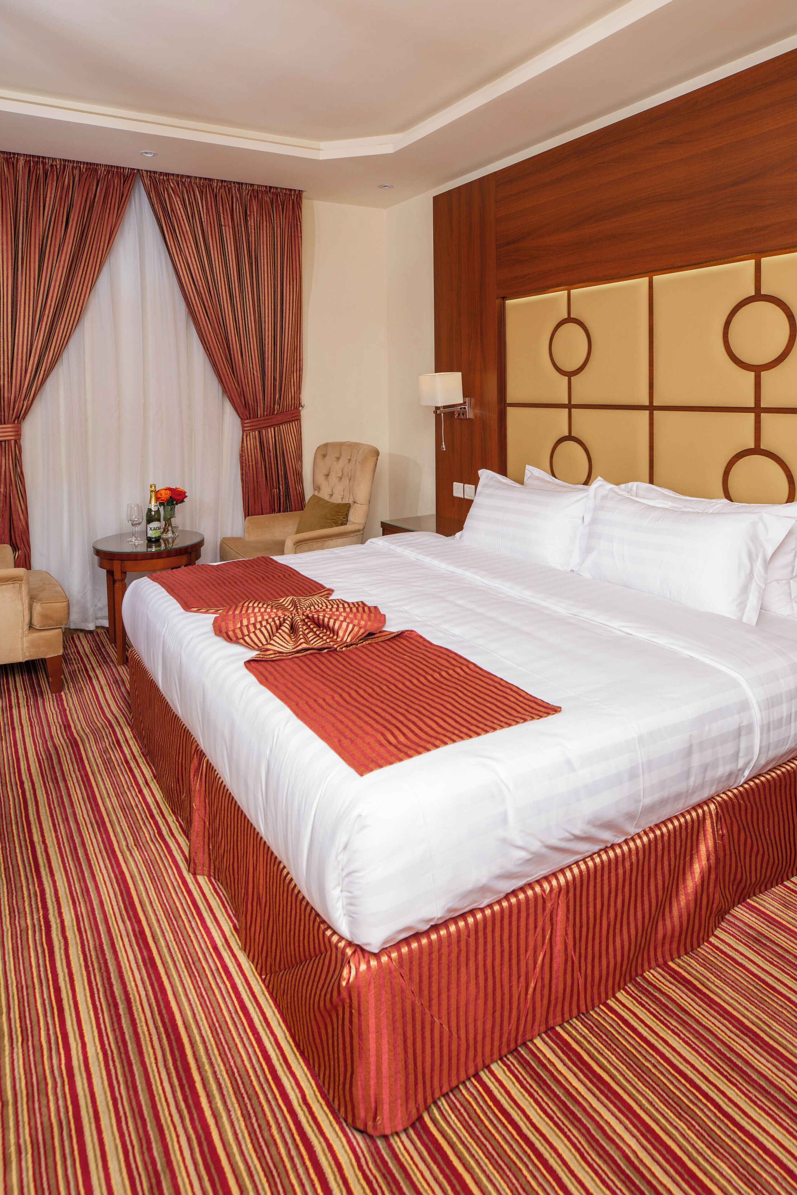 Una habitación de hotel con una cama grande, una zona de estar con una silla y una mesita, y un cabecero de madera.