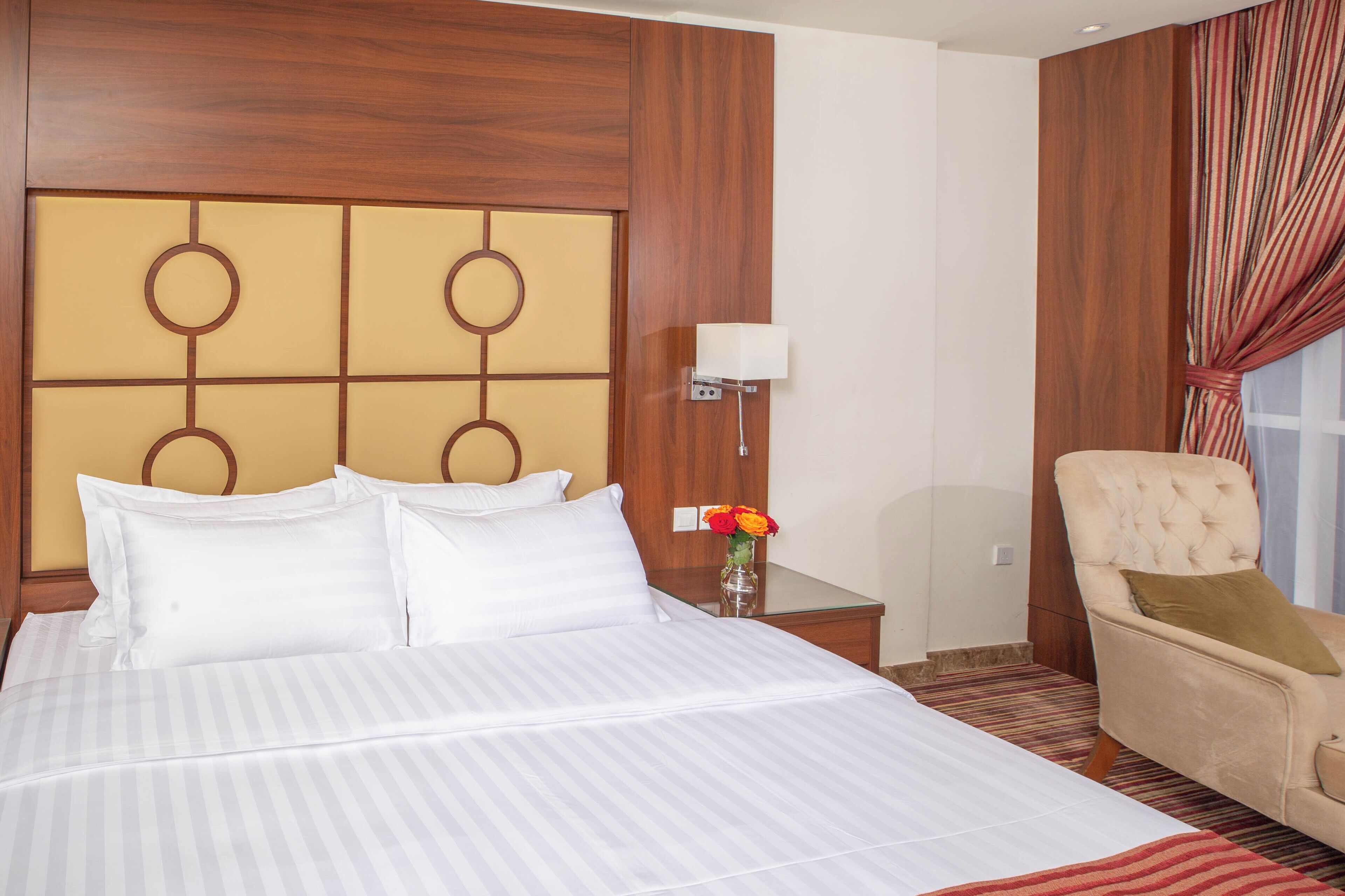 A hotel room with a large bed, a wooden headboard, a bedside table with a lamp, a chair, and a window with curtains.