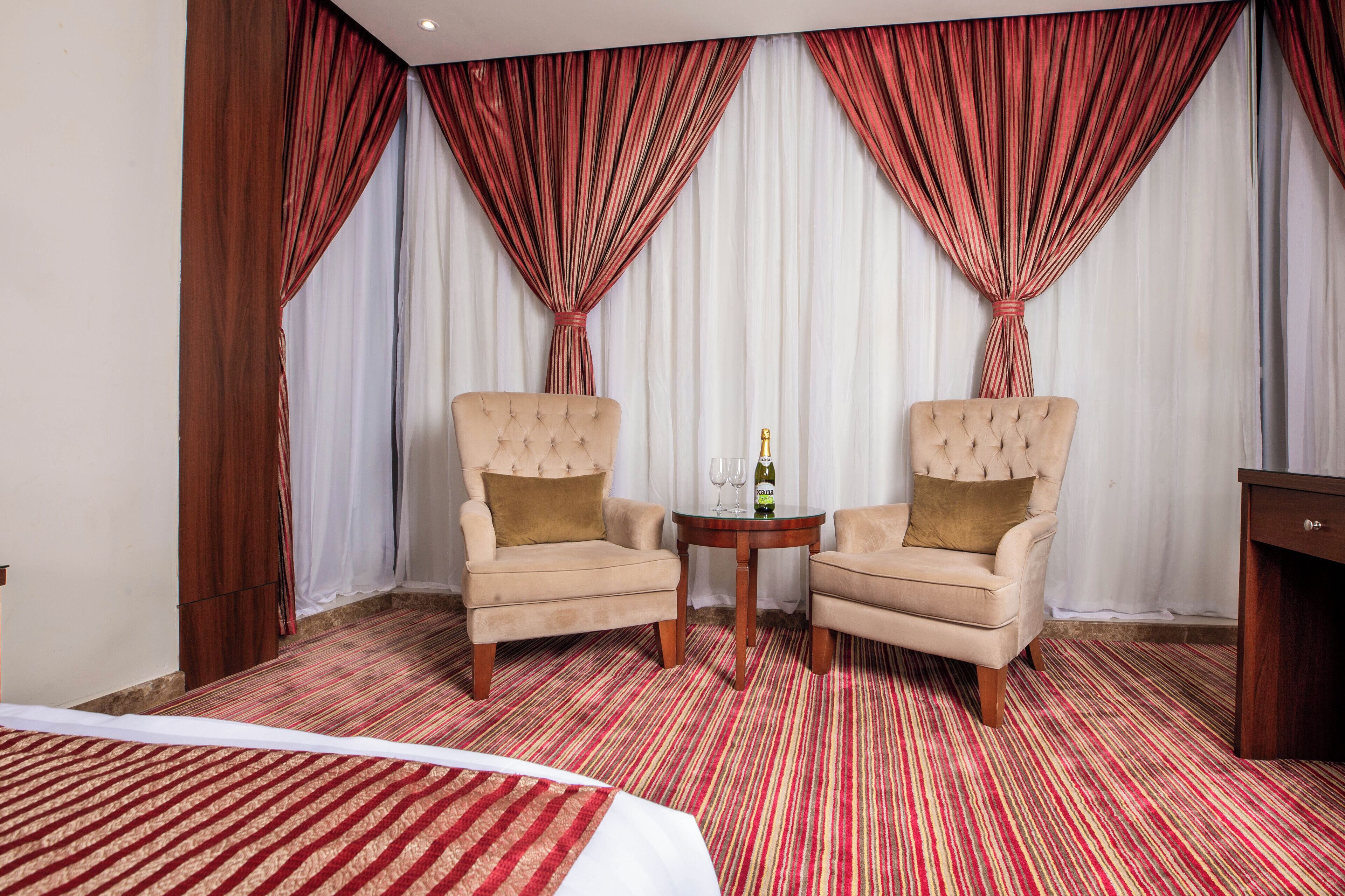 Une chambre d’hôtel avec deux fauteuils, une petite table ronde sur laquelle se trouvent une bouteille et des verres, et un lit recouvert d’une courtepointe à rayures.