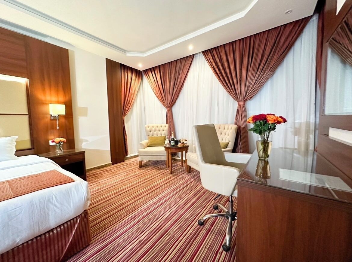 Quarto de hotel com cama, escrivaninha, cadeira, abajur, cortinas e um vaso com flores.