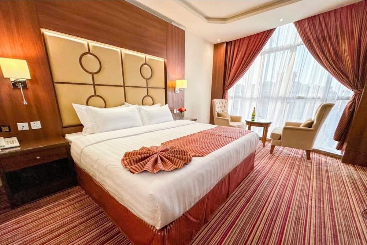 A hotel room with a large bed, a wooden headboard, a bedside table with a lamp, a chair, and a view of the city through large windows.