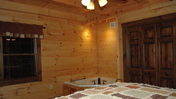 Interior d'una cabana de fusta amb un llit, una finestra amb persianes, una porta de fusta i una banyera.