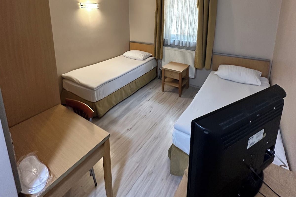 Habitación de hotel con dos camas individuales, un escritorio, una silla, televisión y ventana con cortinas.