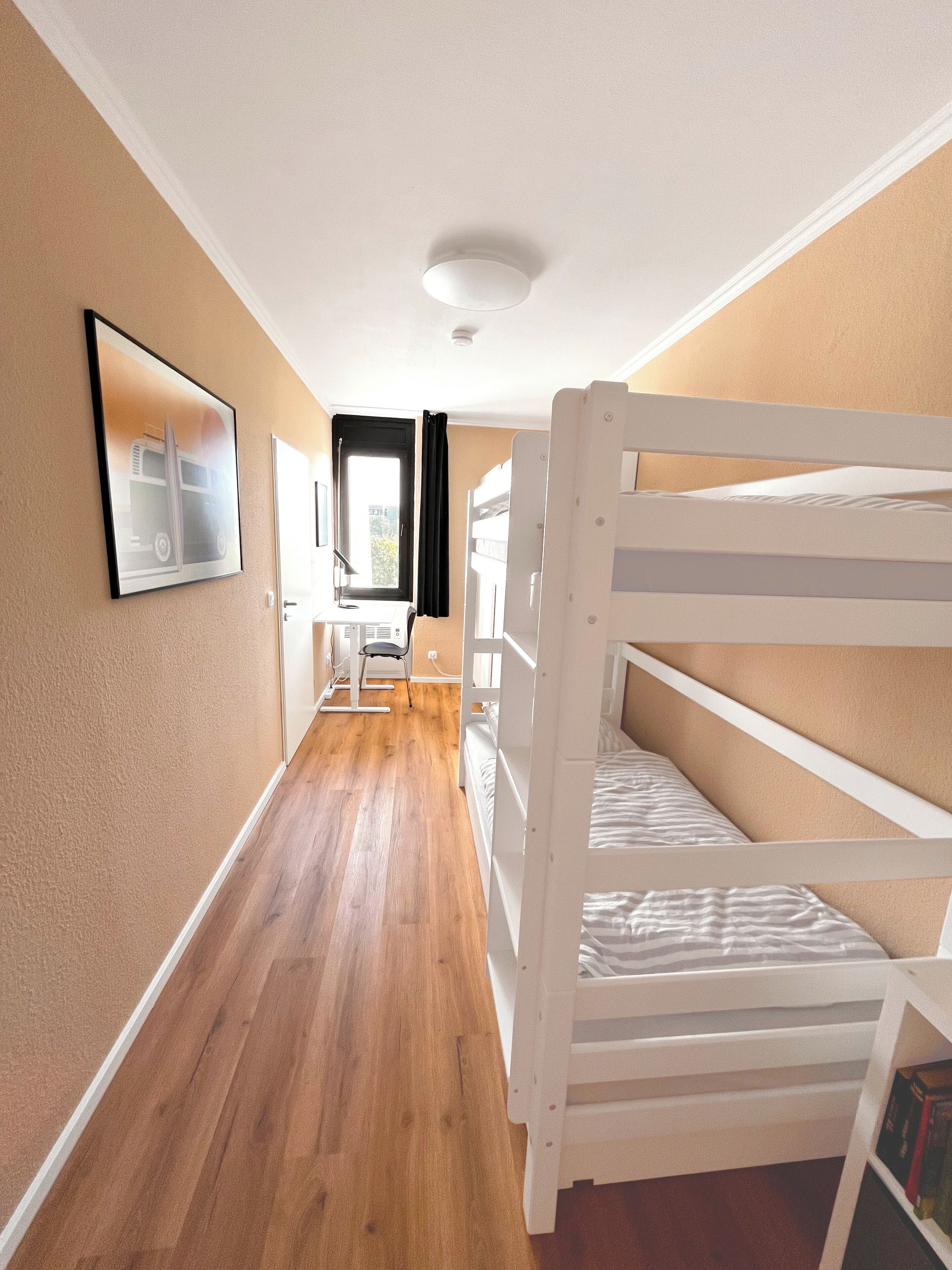 A narrow hallway with wooden flooring, white bunk beds on the right, a framed picture on the left, and a window with curtains in the background.