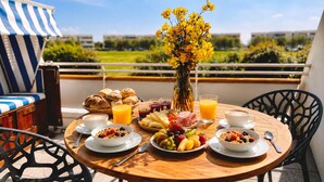 Une table ronde en bois, sur laquelle sont disposés des aliments pour le petit-déjeuner, un vase rempli de fleurs jaunes et une vue sur la verdure et les bâtiments.