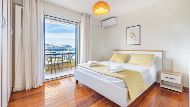 A hotel room with a large bed, a bedside table, a window with curtains, and a view of a cruise ship. - Seaview Portside apartment (Corfu)