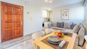 A modern living room with a wooden door, a dining table with chairs, a sofa, and a kitchen in the background. - Seaview Portside apartment (Corfu)
