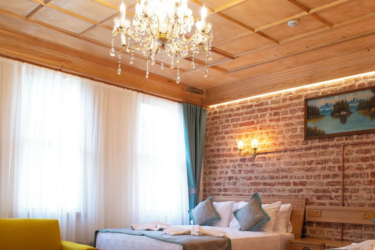 Ein Zimmer mit einem Bett, einem gelben Sessel, einem Holztisch, einer Kristallleuchtre und einer Ziegelwand mit einem Gemälde.