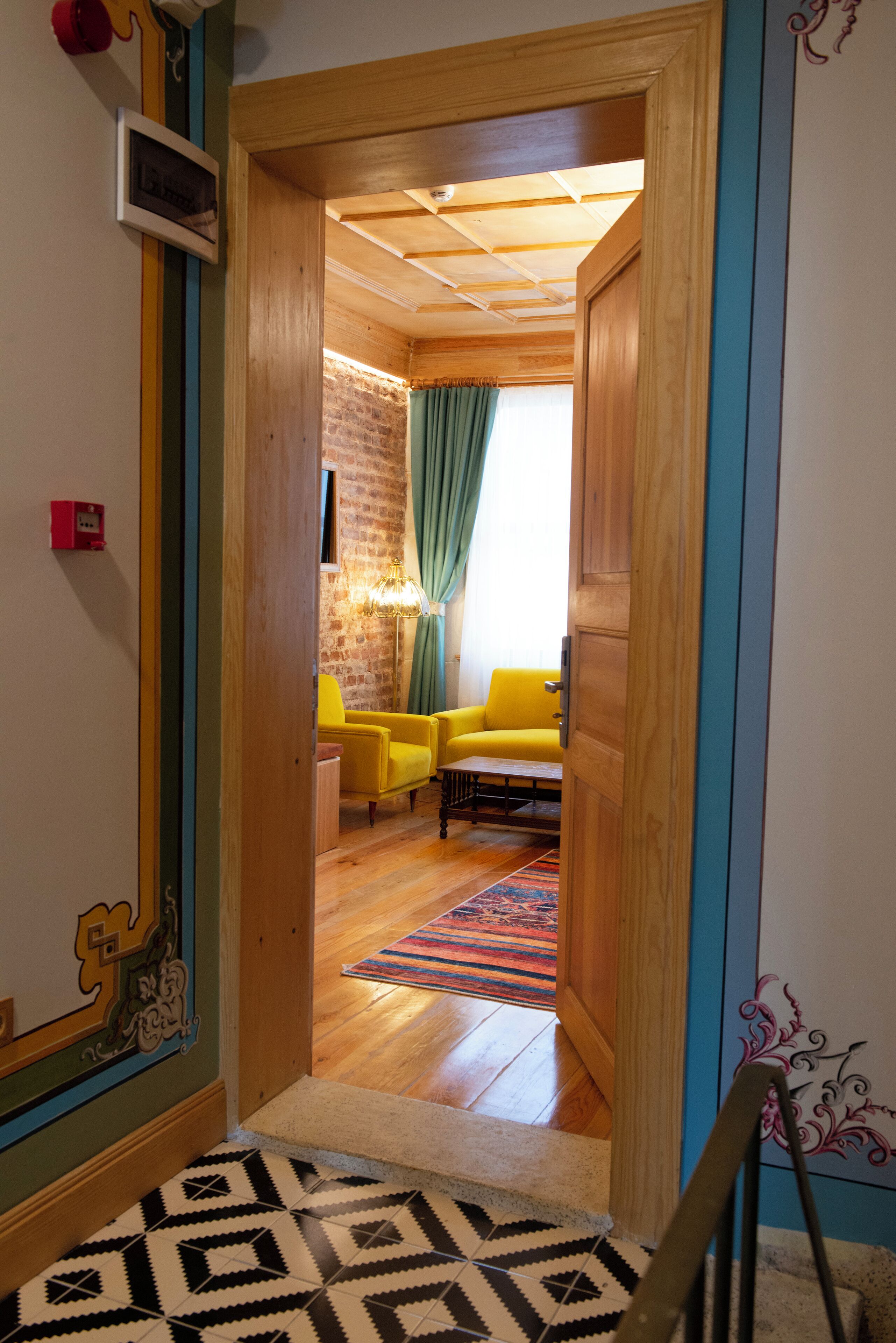 A hallway leading to a room with a brick wall, yellow chairs, a wooden ceiling, and a patterned rug.