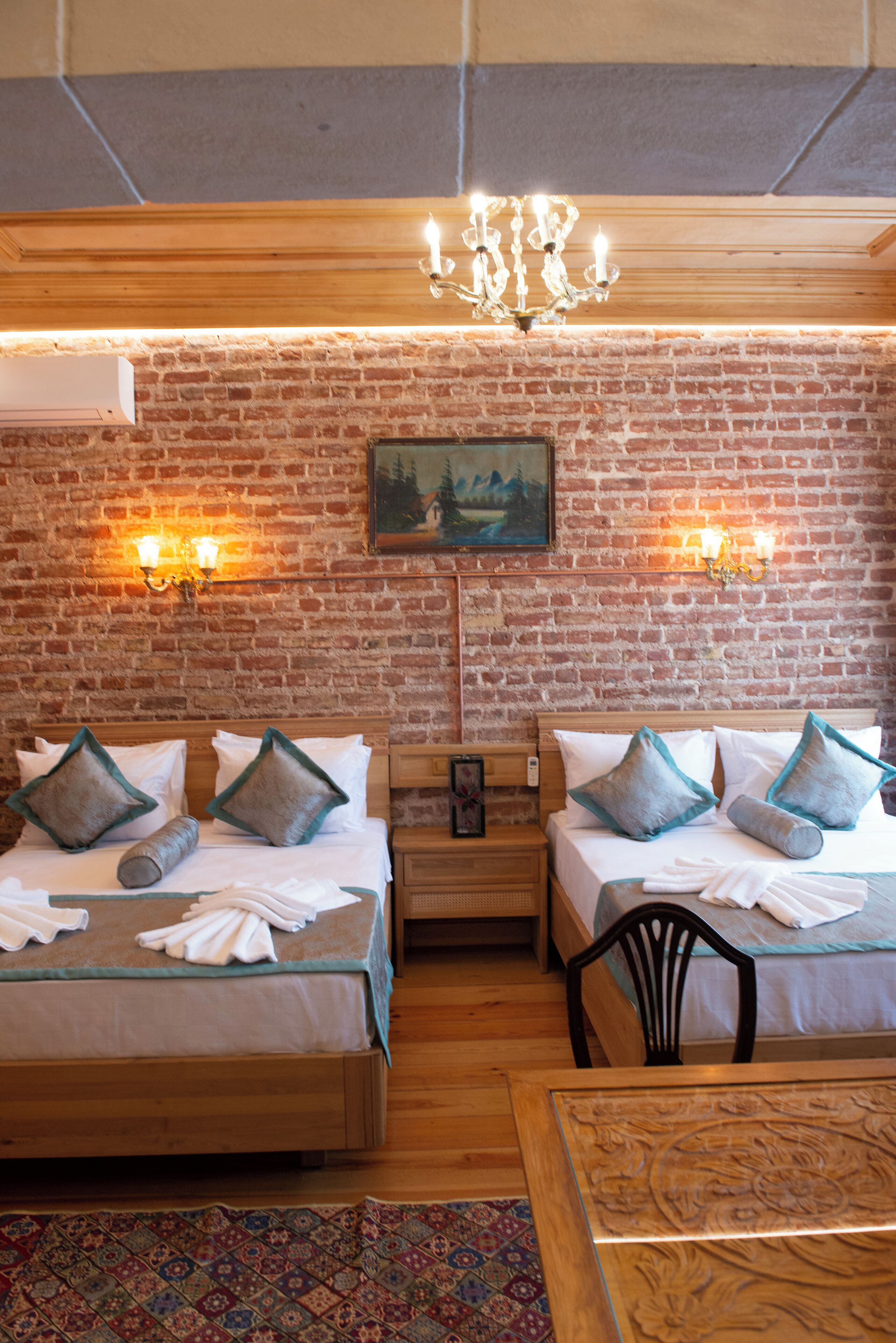 A room with two beds, a wooden table, a chandelier, and a brick wall with a painting.