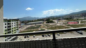 Uitzicht vanaf een balkon over een parkeerplaats met meerdere auto's, een gebouw met een groen en wit gevel en een bergachtig landschap op de achtergrond.