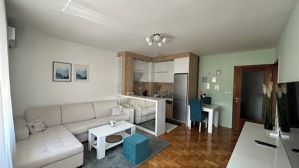 Een moderne woonkamer met een bank, salontafel en poef, een keuken met een koelkast en fornuis, en een eetgedeelte met een tafel en stoelen.