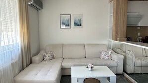 Een moderne woonkamer met een hoekbank, een witte salontafel en een blauwe poef. Twee ingelijste foto's hangen aan de muur, er is een keuken met een magnetron en een groot raam met dunne gordijnen.