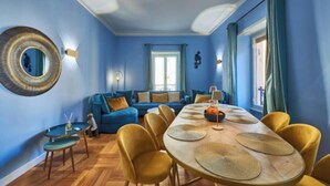 A room with a blue wall, a round mirror, a sofa, a dining table with yellow chairs, and a small table with a lamp. - A Blend of Historical Elegance and Modern Comfort (Roma)