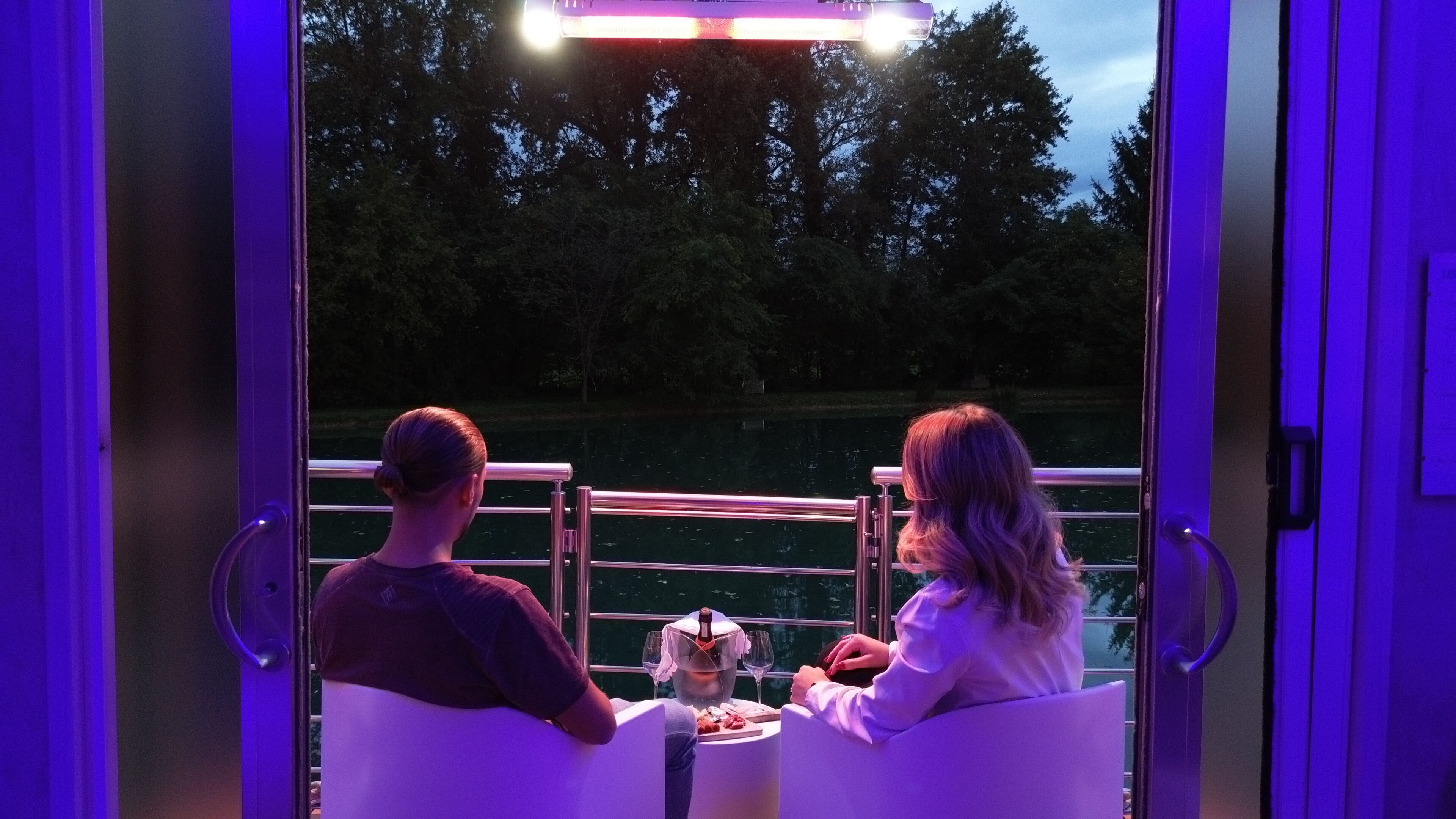 Una pareja sentada en una mesa con vista a un lago y árboles, en una habitación con iluminación morada.