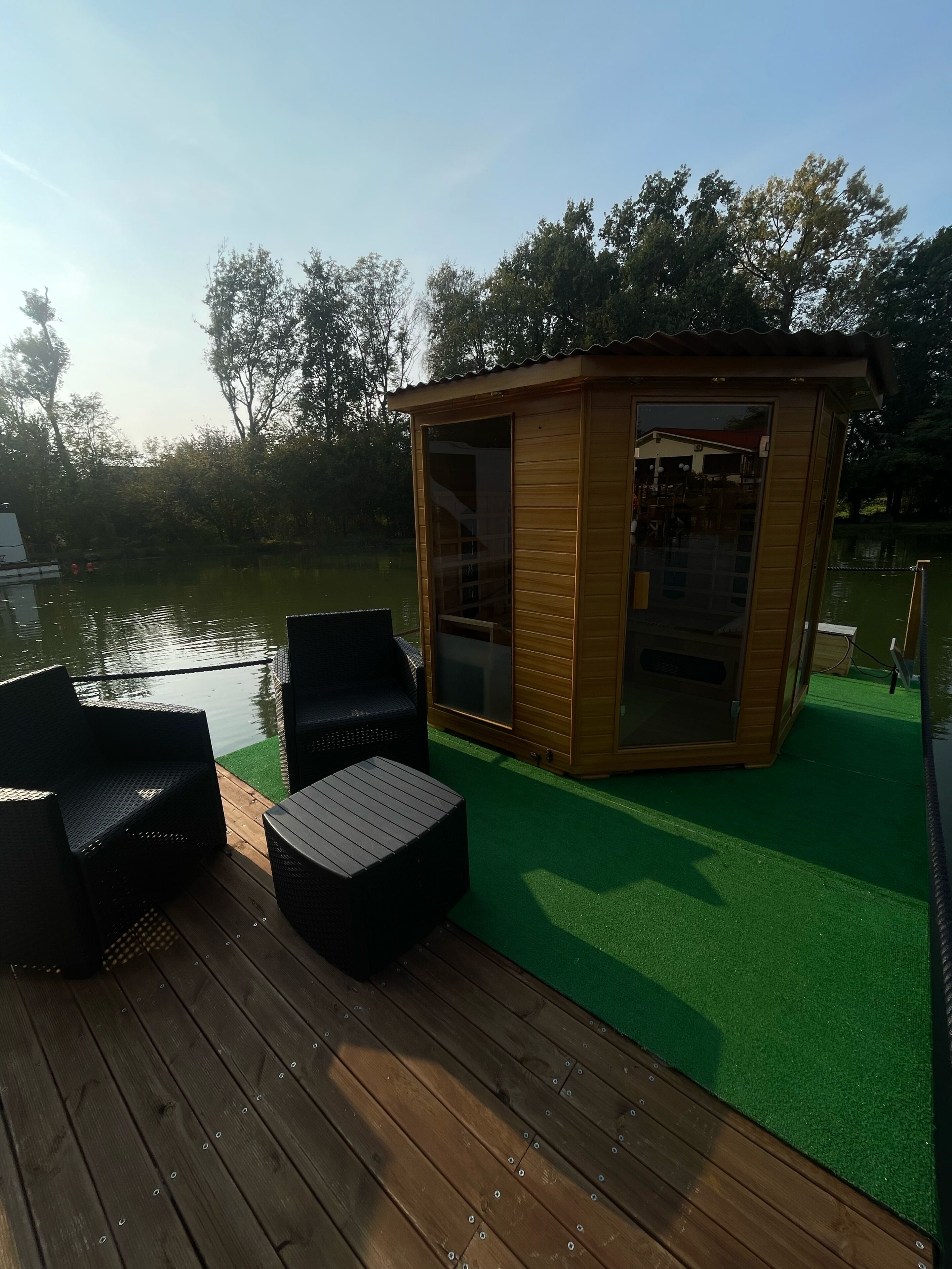 Eine Holzterrasse mit zwei schwarzen Korbstühlen und einem kleinen Tisch, einer hölzernen Struktur mit Glasverkleidung und einer Fläche mit grüner Kunstrasen.