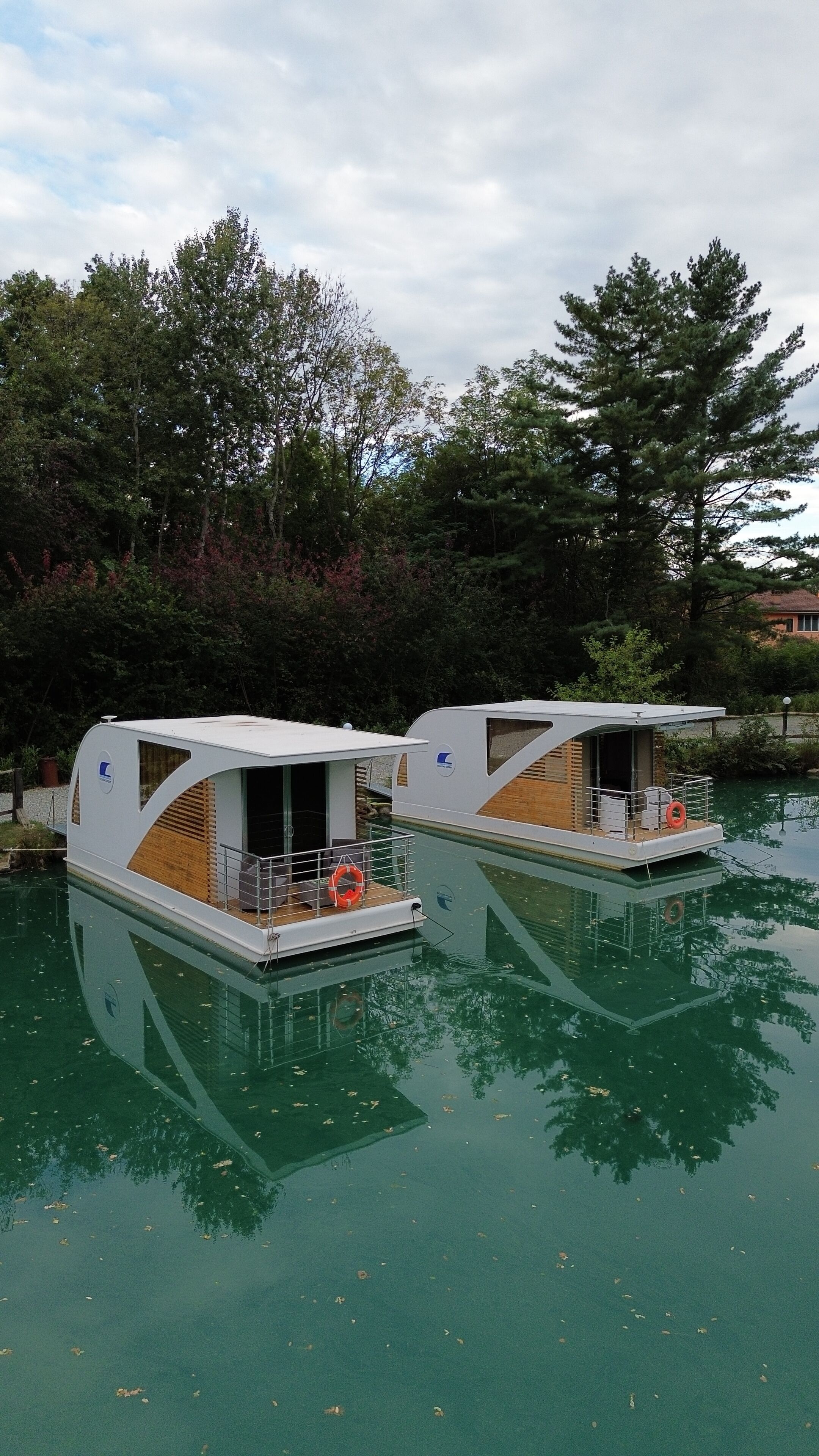 Two floating houseboats with open decks and seating areas, surrounded by calm water and trees.