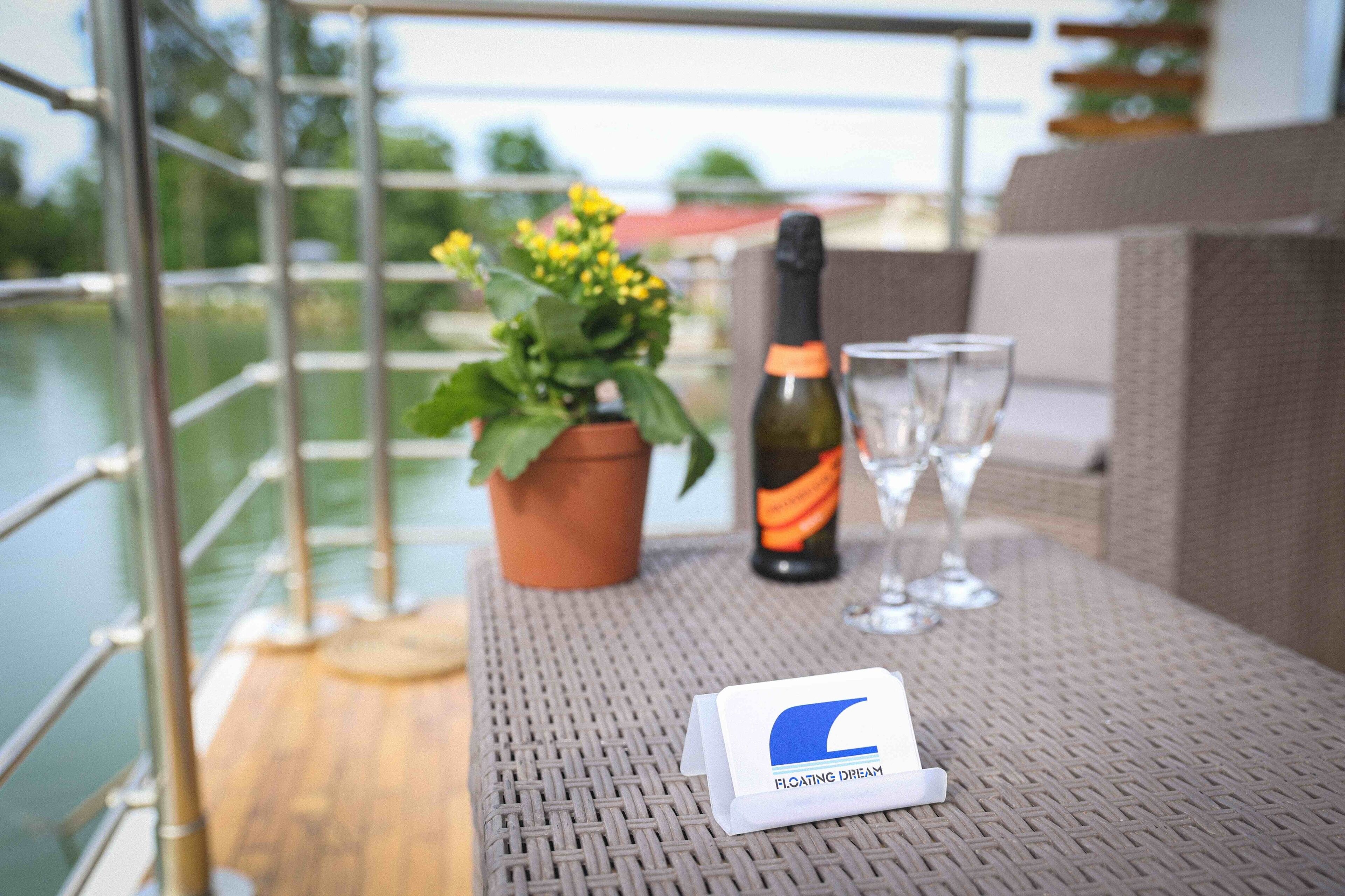 Ein Tisch mit einer Flasche, Gläsern und einer Pflanze auf einem Balkon mit Blick auf ein Gewässer.