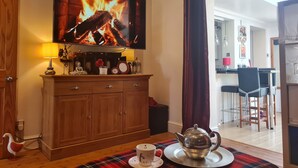 Een gezellige woonkamer met een televisie waarop een haardbeeld te zien is, een houten kast, een tafel met een theepot en kopje, en een eethoek op de achtergrond.