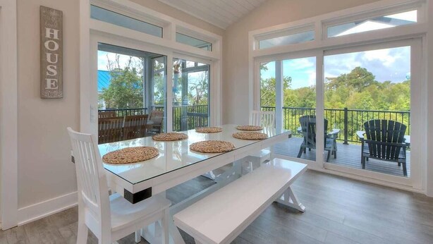 A dining area with a glass table, white chairs, and a bench. There are large windows and a balcony with outdoor seating. - Slow n Steady: Swim Spa, Elevator, Beach Walk (Oak Island)