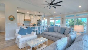A modern living room with a white sofa, grey sofa, and a glass coffee table. There is a kitchen area with white cabinets and a dining table with chairs. - Slow n Steady: Swim Spa, Elevator, Beach Walk (Oak Island)