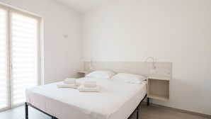 A minimalist hotel room with a bed, two bedside tables, and a large window with blinds. - Das Halte Haus - Room 302 (Olbia)