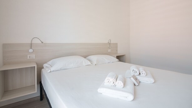 A neatly made bed with white linens and towels, a wooden headboard, and a small bedside table with a lamp. - Das Halte Haus - Room 302 (Olbia)