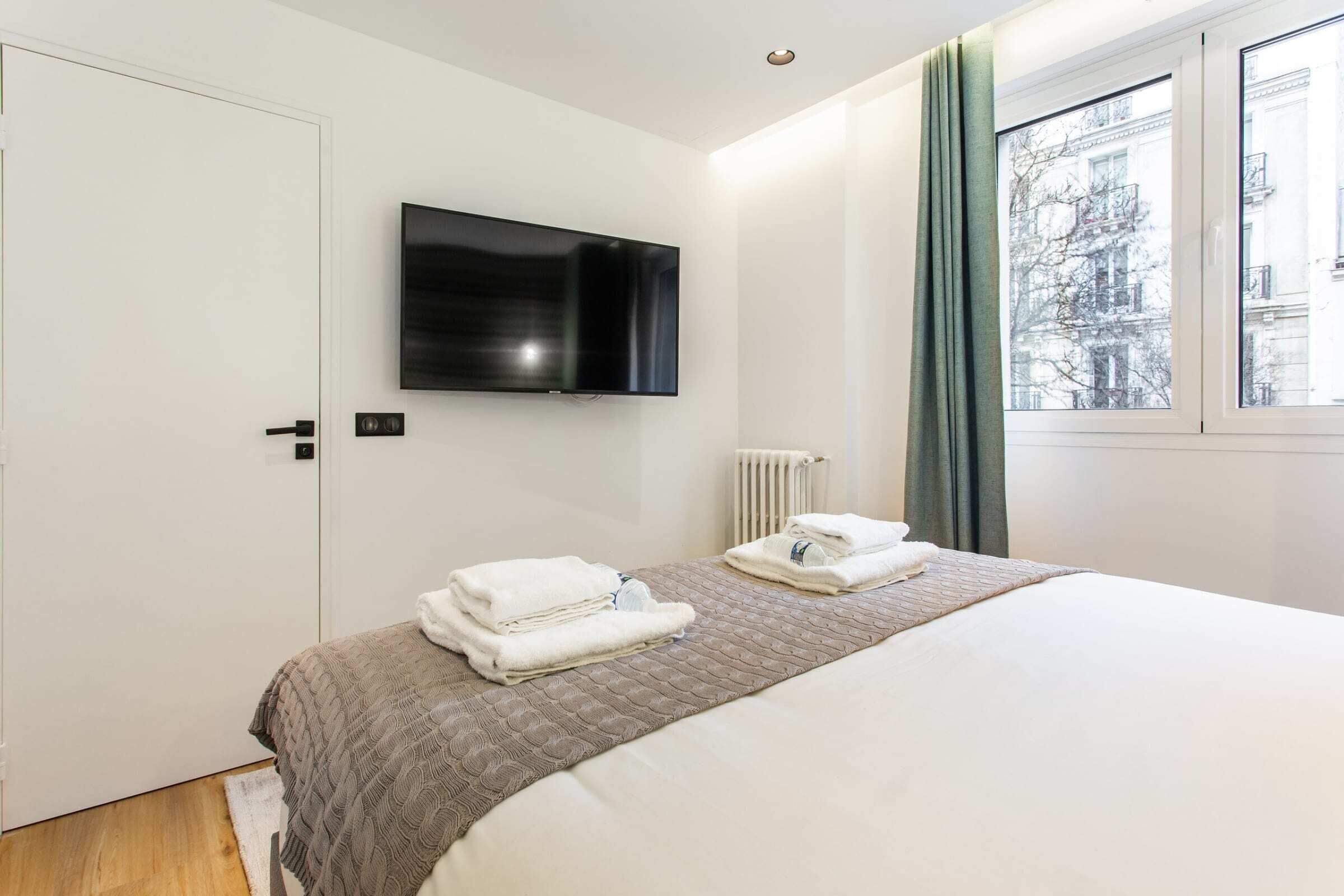 Hotelkamer met een bed, een aan de muur bevestigde televisie, een radiator en een raam met gordijnen.
